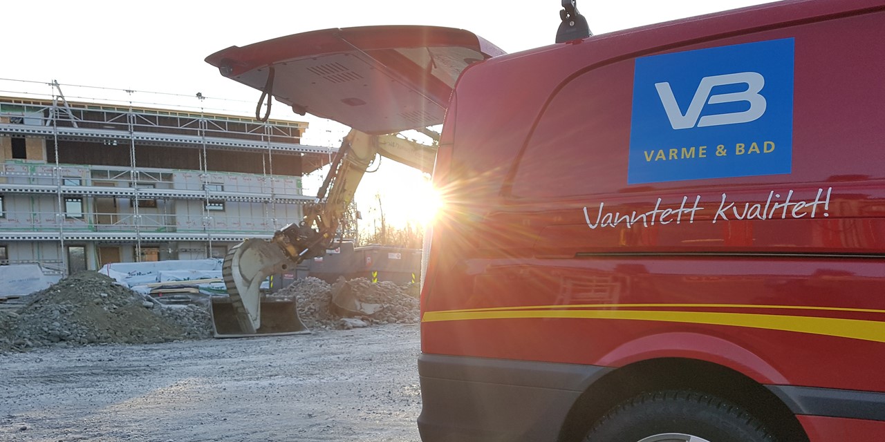 Rød varebil fra VB i motlys fra lav sol og foran byggeplass med stillas og gravemaskin.